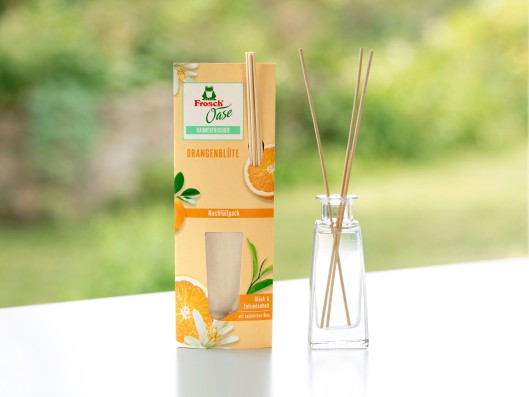 A room fragrance glass bottle with reed diffusers stands next to a Frosch Oase Orange scent sachet