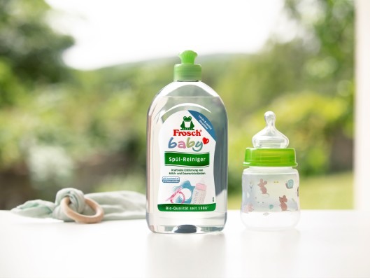 A Frosch baby dishwashing liquid bottle stands next to a baby bottle in front of a window