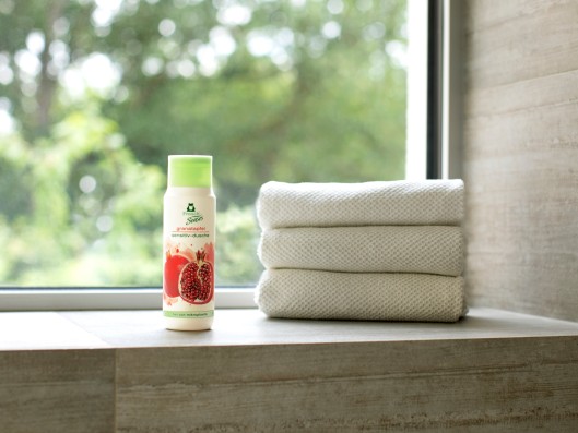 Im Badezimmer vor einem Fenster steht eine Flasche Frosch Sensitiv-Dusche Granatapfel neben gestapelten Handtüchern