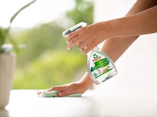 A plain surface on which a potted plant stands is sprayed with a spray bottle of Frosch Kitchen Hygiene Cleaner and wiped with a cloth