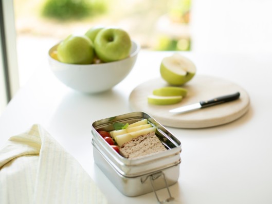 A lunch box, a bowl with apples and a cutting board with a knife on a white table