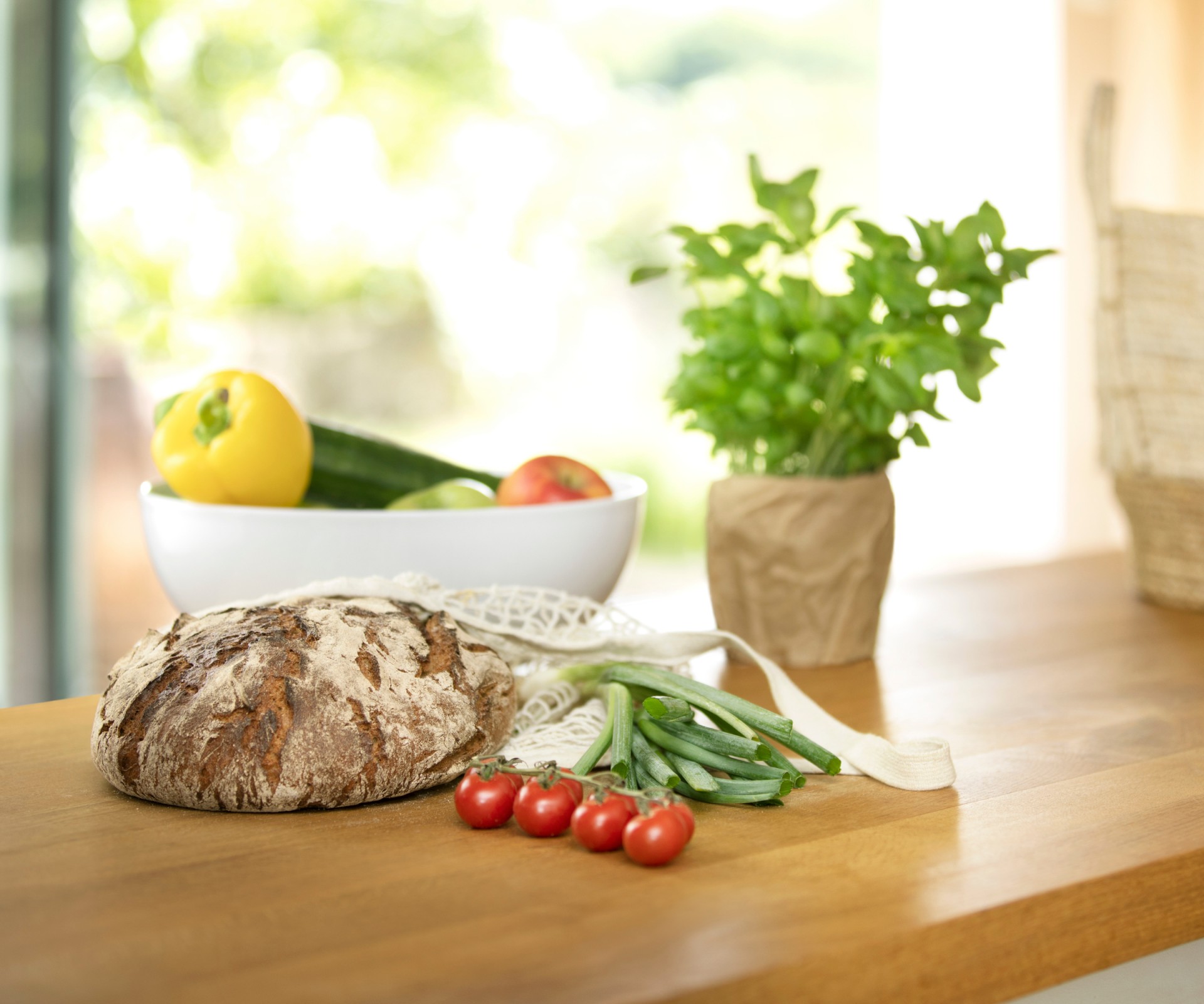 Auf einem Holztisch liegen ein Brot, Tomaten, Basilikum und eine Schale mit Gemüse