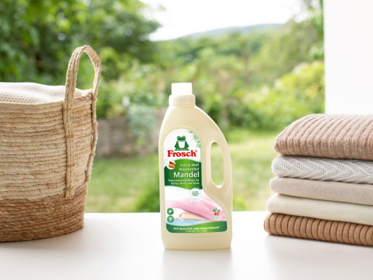 A Frosch Delicate almond detergent pouch stands between a basket and stacked towels
